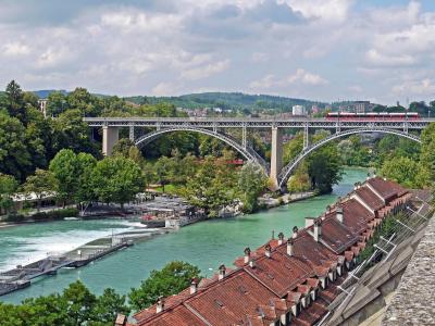 Panoramic view of the historic Kirchenfeld Bridge over the Aare River in Bern, surrounded by lush greenery and traditional rooftops.