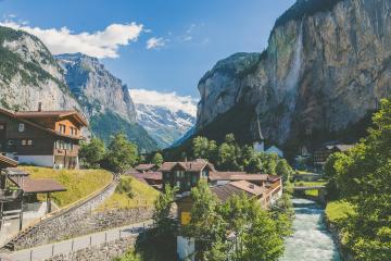 A scenic view of a Swiss village nestled in a valley between steep mountains and a waterfall, featuring traditional chalets and a river.