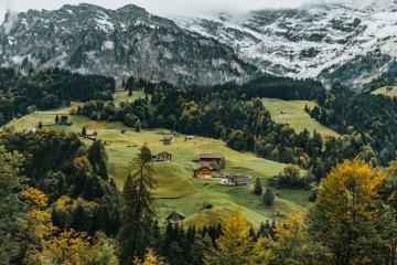 A scenic landscape view of rolling green hills dotted with small wooden chalets and surrounded by dense forests, with snow-capped mountains in the background under an overcast sky.