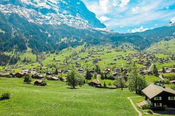 A scenic view of a village with traditional chalets nestled in a green valley, backed by a large, snow-dusted mountain peak under a blue sky.