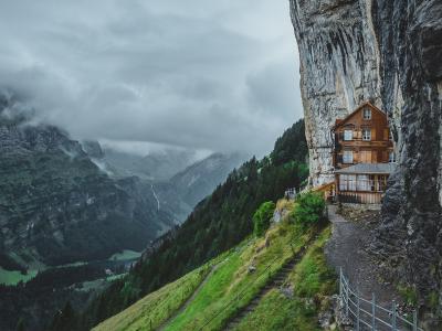 A wooden chalet-style building built into the vertical face of a limestone cliff, with a metal walkway and railing leading to it, set against a backdrop of green alpine slopes and cloudy skies.