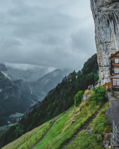 A wooden chalet-style building built into the vertical face of a limestone cliff, with a metal walkway and railing leading to it, set against a backdrop of green alpine slopes and cloudy skies.