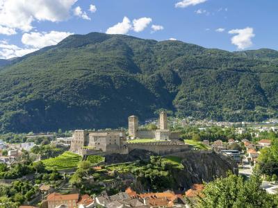 A high-angle daytime view of the Castelgrande fortress and its two towers perched on a rocky promontory, overlooking the town of Bellinzona with a large green mountain in the background.