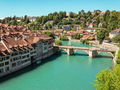 A view of the Aare River in Bern, Switzerland, featuring the historic Nydeggbrücke bridge and the distinctive red-tiled roofs of the Old Town buildings along the riverbank.