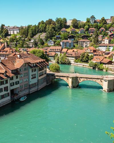 A view of the Aare River in Bern, Switzerland, featuring the historic Nydeggbrücke bridge and the distinctive red-tiled roofs of the Old Town buildings along the riverbank.