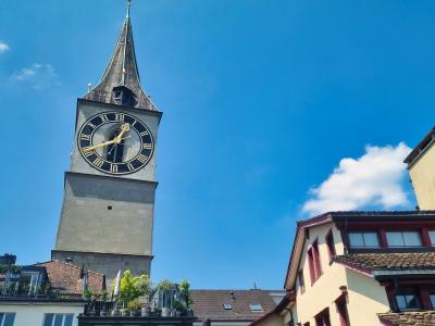 A tall stone church tower with a large clock face and a spire, set against a blue sky, with other buildings in the foreground.