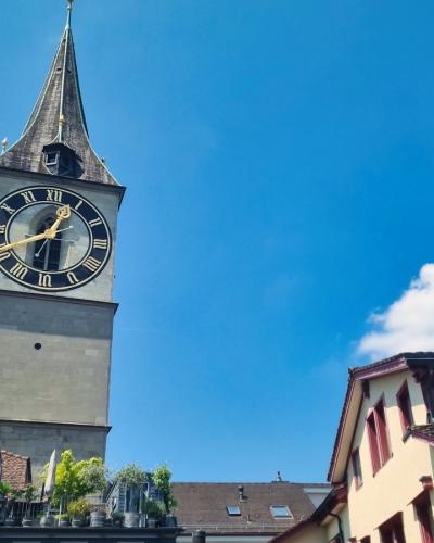 A tall stone church tower with a large clock face and a spire, set against a blue sky, with other buildings in the foreground.