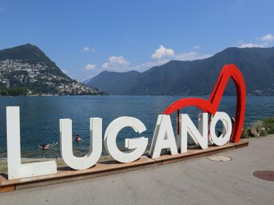 A large outdoor sign spelling "LUGANO" in white letters with a red heart symbol, situated on a paved area next to a lake with mountains in the background.