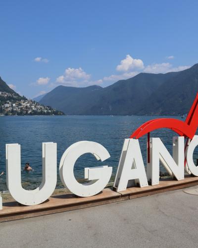 A large outdoor sign spelling "LUGANO" in white letters with a red heart symbol, situated on a paved area next to a lake with mountains in the background.