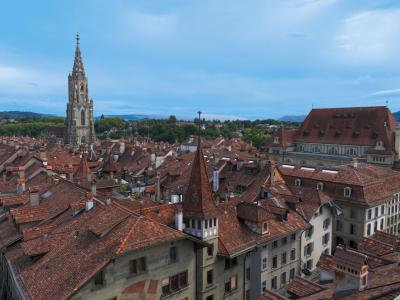 A high-angle view of the red-tiled rooftops of Bern's Old City, dominated by the Gothic spire of the Bern Minster on the left and the Zytglogge clock tower in the foreground.