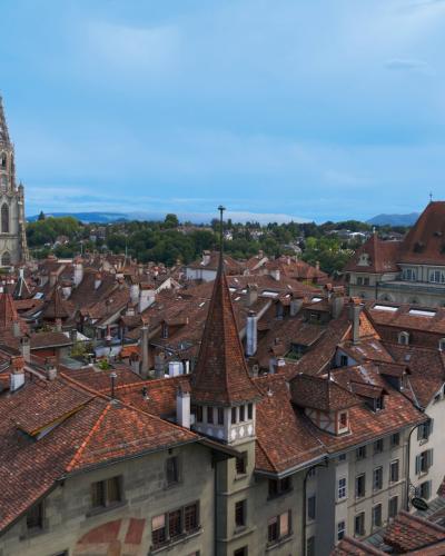 A high-angle view of the red-tiled rooftops of Bern's Old City, dominated by the Gothic spire of the Bern Minster on the left and the Zytglogge clock tower in the foreground.