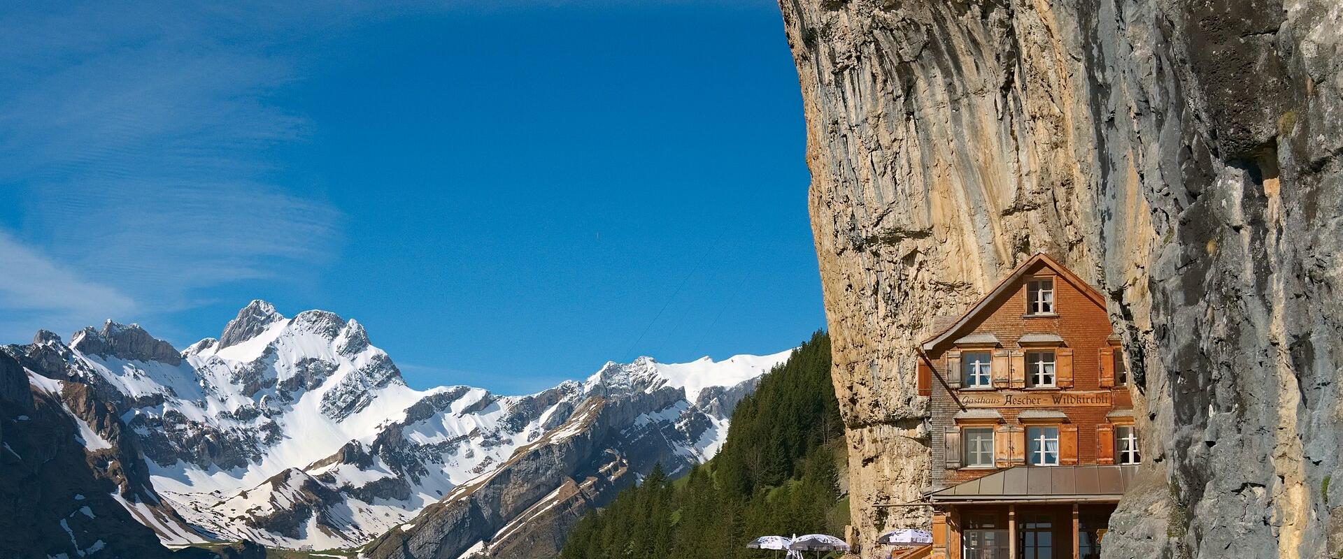 A wooden building built into a vertical rock face with a sign reading "Aescher Wildkirchli" above the windows, set against a backdrop of snow-capped mountains.