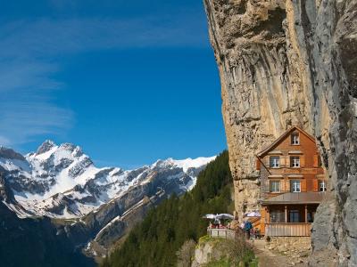 A wooden building built into a vertical rock face with a sign reading "Aescher Wildkirchli" above the windows, set against a backdrop of snow-capped mountains.