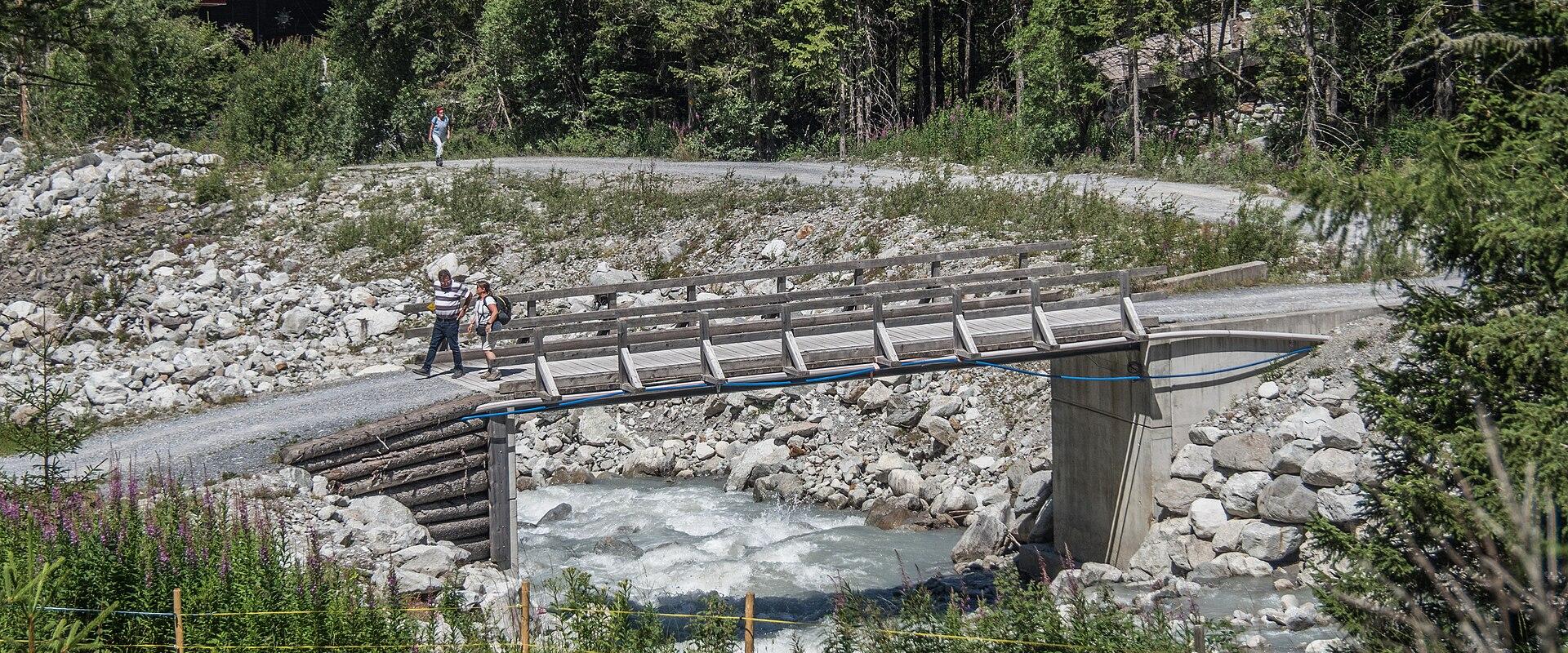 A wooden footbridge crossing a rocky riverbed with a gravel road and hikers in the background, set against a dense forest.