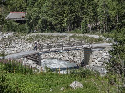 A wooden footbridge crossing a rocky riverbed with a gravel road and hikers in the background, set against a dense forest.
