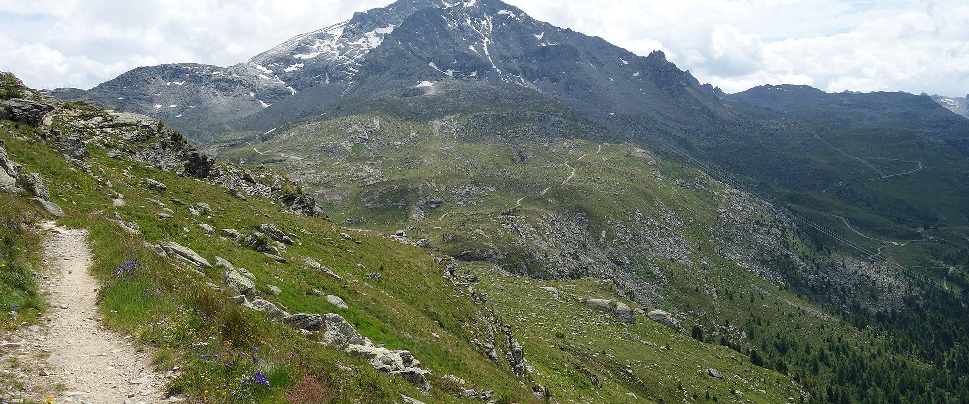 A dirt hiking trail winds up a grassy alpine slope in the foreground, leading towards a large, rocky mountain peak with patches of snow in the background under a cloudy sky.