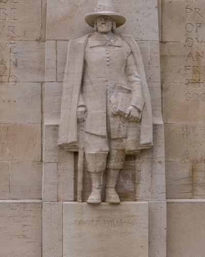A stone relief sculpture of a man in 17th-century attire holding a book, with the name "ROGER WILLIAMS" inscribed on the base, set within a larger stone wall featuring other inscriptions.