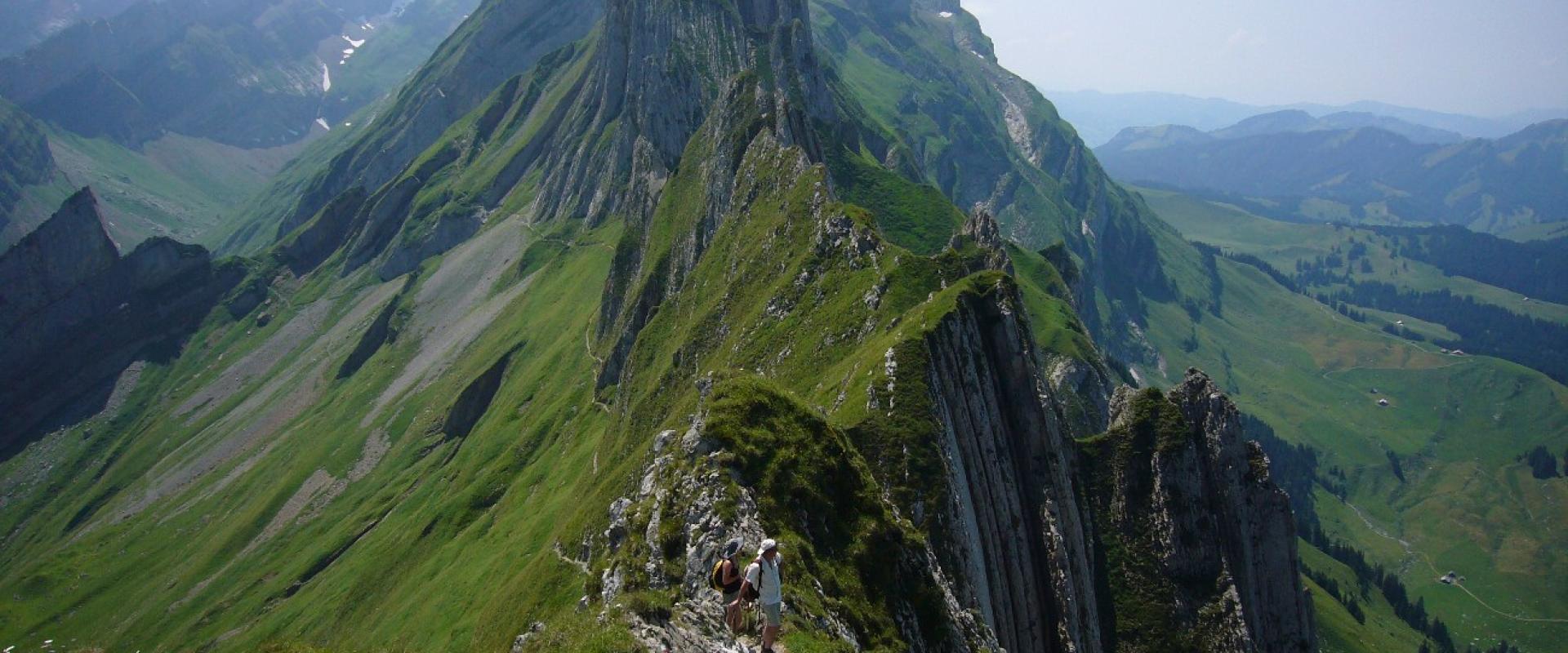 A narrow, rocky mountain ridge with hikers walking along a trail, set against a backdrop of green alpine slopes and distant peaks under a blue sky.