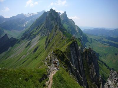 A narrow, rocky mountain ridge with hikers walking along a trail, set against a backdrop of green alpine slopes and distant peaks under a blue sky.