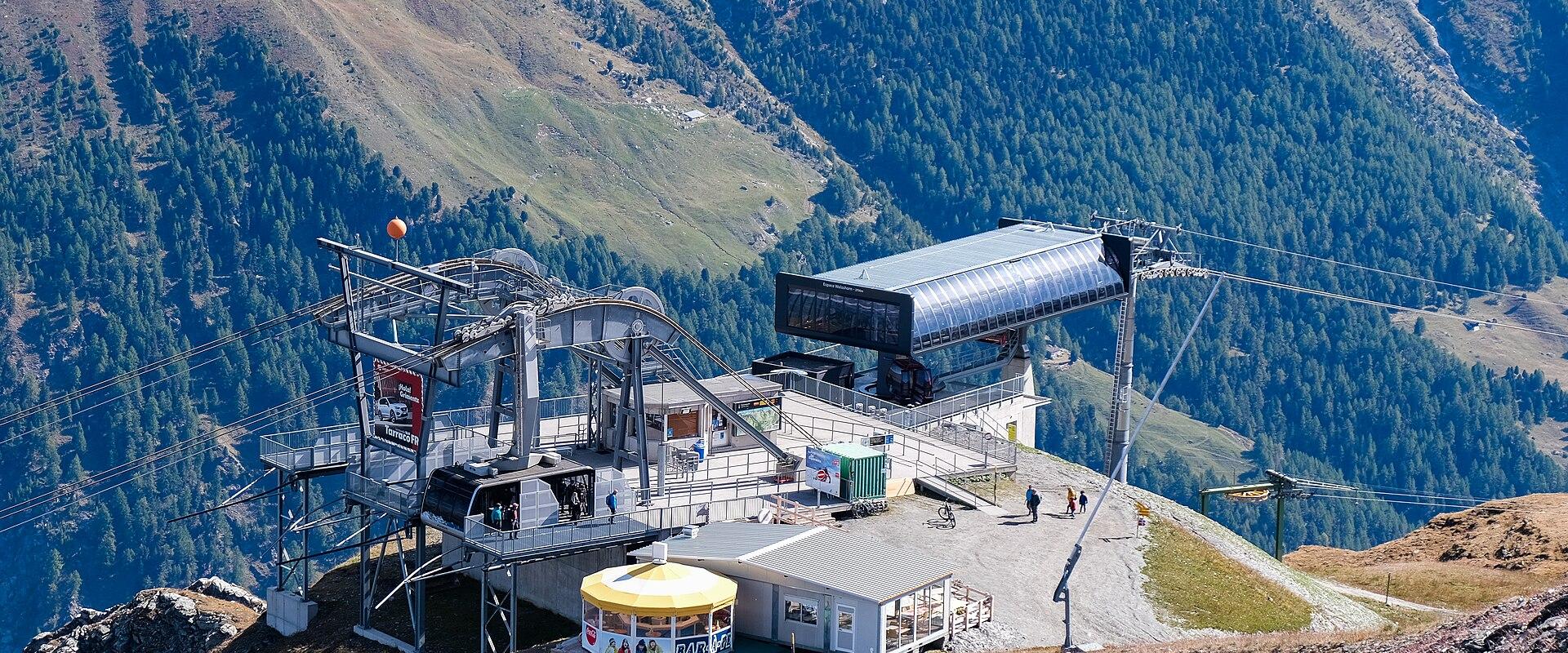 A high-altitude ski lift station with a yellow kiosk and metal infrastructure situated on a rocky ridge, overlooking a steep, forested valley.