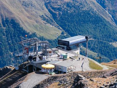 A high-altitude ski lift station with a yellow kiosk and metal infrastructure situated on a rocky ridge, overlooking a steep, forested valley.