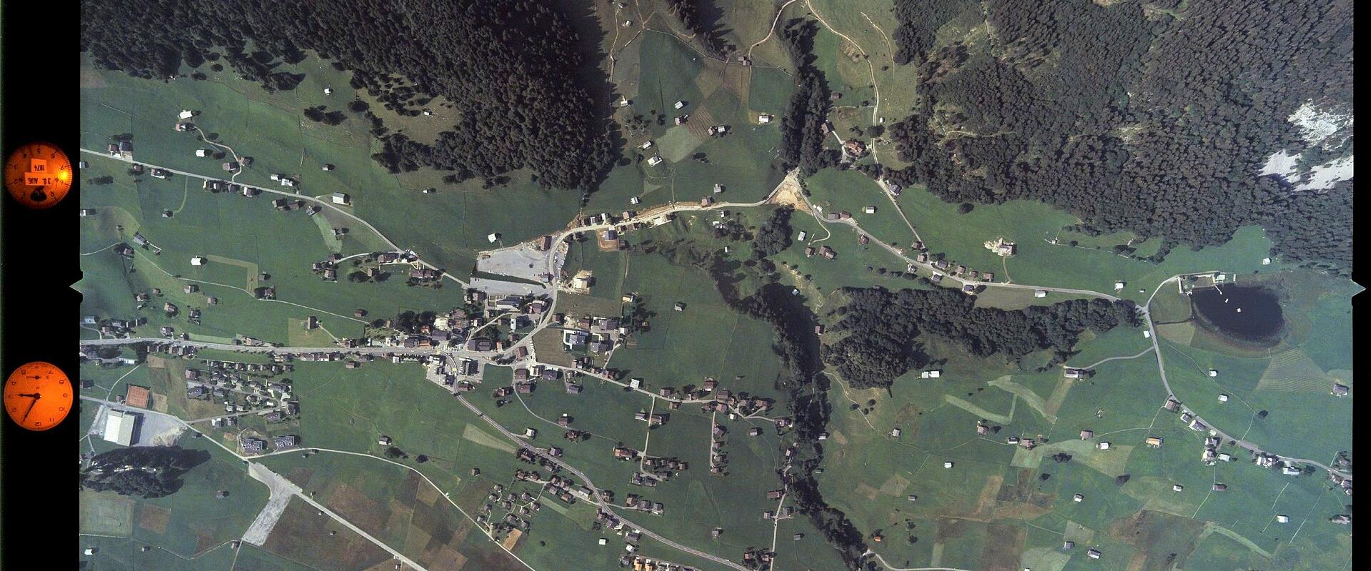 An aerial photograph showing a rural valley settlement with scattered houses, fields, and forested hillsides, featuring orange color correction markers along the left edge.