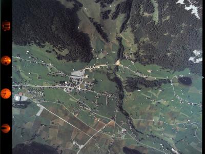 An aerial photograph showing a rural valley settlement with scattered houses, fields, and forested hillsides, featuring orange color correction markers along the left edge.