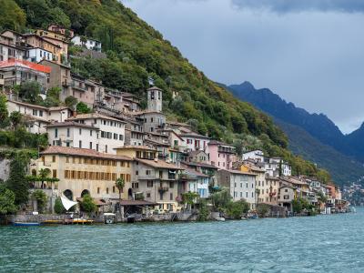 A view of a densely built village with colorful houses and a stone church tower climbing a steep, forested hillside directly adjacent to a large lake, with mountains in the background under a cloudy sky.