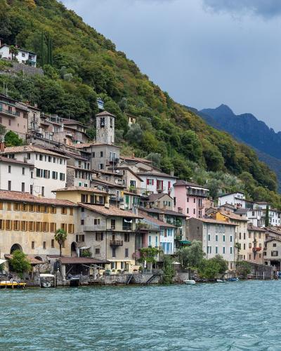 A view of a densely built village with colorful houses and a stone church tower climbing a steep, forested hillside directly adjacent to a large lake, with mountains in the background under a cloudy sky.