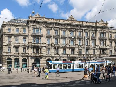 A wide-angle street view of a grand, ornate Beaux-Arts building with “CREDIT SUISSE” signage on its roof, featuring arched ground-floor windows, multiple balconies, and classical statuary; in front, a blue-and-white tram passes along tracks, with