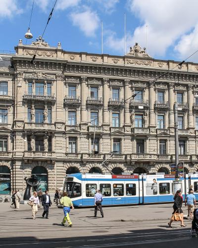 A wide-angle street view of a grand, ornate Beaux-Arts building with “CREDIT SUISSE” signage on its roof, featuring arched ground-floor windows, multiple balconies, and classical statuary; in front, a blue-and-white tram passes along tracks, with