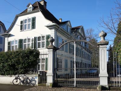 A white, multi-story manor house with green shutters and a decorative wrought-iron gate, featuring house numbers 61 and 65 on the pillars.