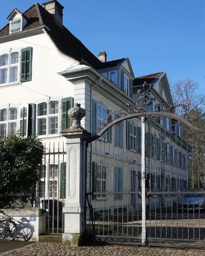 A white, multi-story manor house with green shutters and a decorative wrought-iron gate, featuring house numbers 61 and 65 on the pillars.