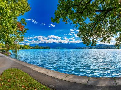 A paved promenade curves along the edge of a large, bright blue lake, framed by green trees in the foreground with distant mountains visible across the water under a blue sky.