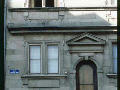 A close-up of a stone building facade featuring a secondary entrance with an arched wooden door, two windows with white curtains, and a blue street sign reading "Rue de l'HOTEL-DE-VILLE".