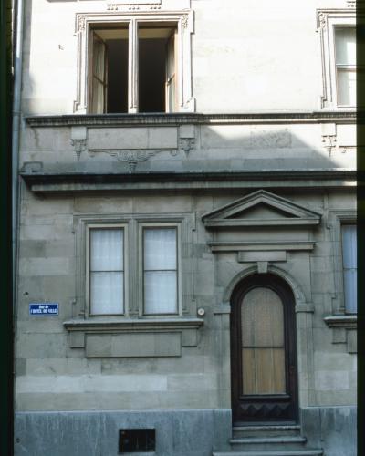 A close-up of a stone building facade featuring a secondary entrance with an arched wooden door, two windows with white curtains, and a blue street sign reading "Rue de l'HOTEL-DE-VILLE".