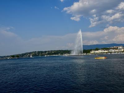 A tall water fountain shooting a plume into the air from a lake, with a shoreline of buildings and hills in the background under a partly cloudy sky.