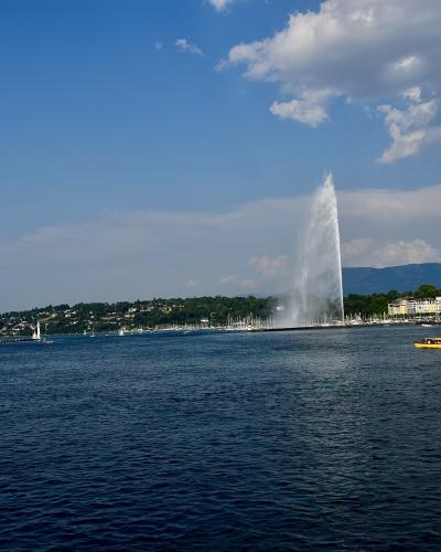 A tall water fountain shooting a plume into the air from a lake, with a shoreline of buildings and hills in the background under a partly cloudy sky.