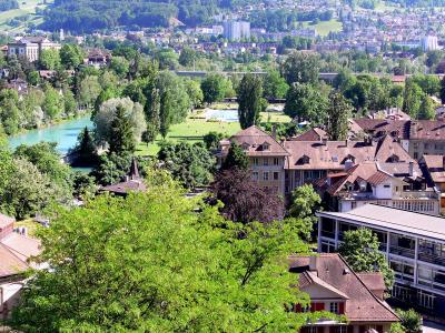 A high-angle view of the Aare river in Bern, Switzerland, showing the Freibad Marzili swimming area with its pools and grassy banks, surrounded by trees and residential buildings, with the city and hills in the background.