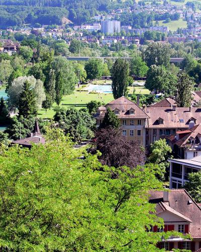A high-angle view of the Aare river in Bern, Switzerland, showing the Freibad Marzili swimming area with its pools and grassy banks, surrounded by trees and residential buildings, with the city and hills in the background.