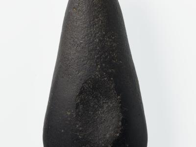 A studio photograph of a dark, polished stone axe head (stone adze) against a plain white background.