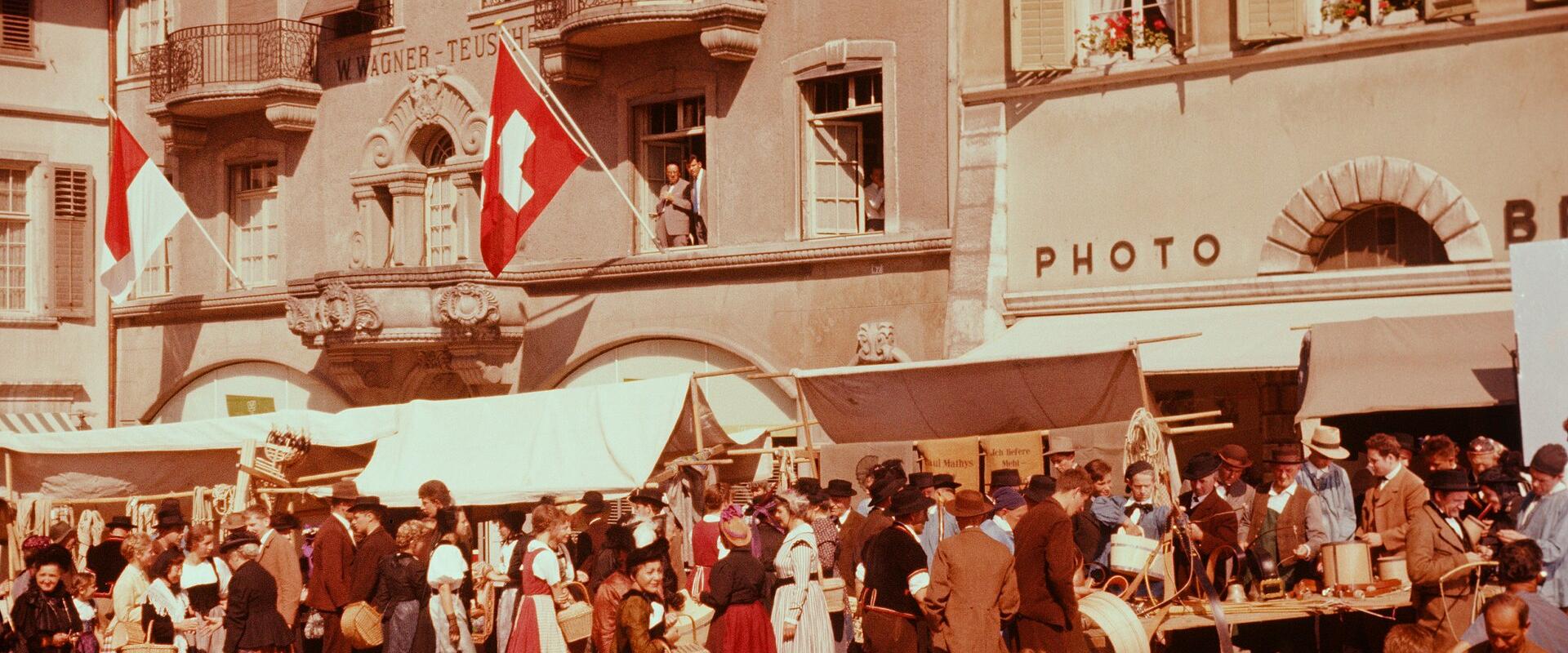 A crowded outdoor market scene in a European town square with people in traditional dress, market stalls, and buildings featuring Swiss flags and signage reading "SOLOTHURN" and "WAGNER-TEUSCH".
