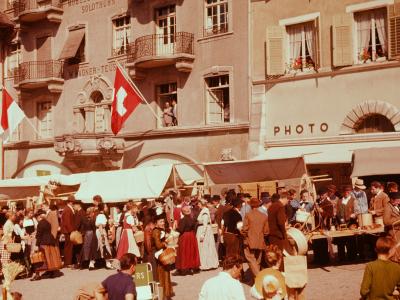 A crowded outdoor market scene in a European town square with people in traditional dress, market stalls, and buildings featuring Swiss flags and signage reading "SOLOTHURN" and "WAGNER-TEUSCH".