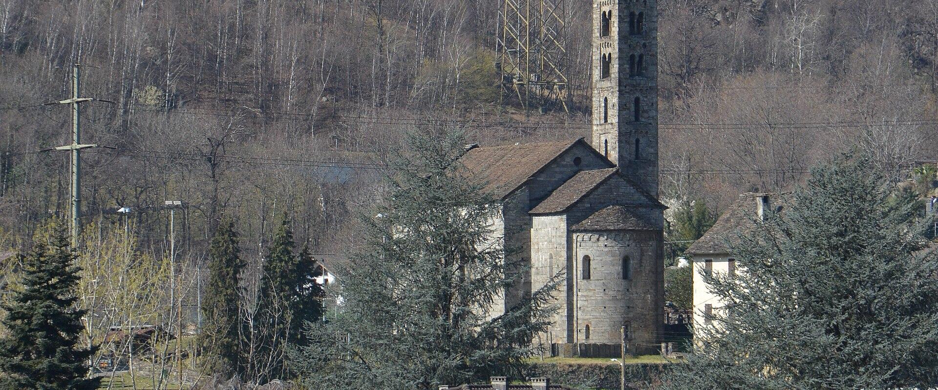 A stone church with a tall, square bell tower and a rounded apse, situated on a hillside behind modern residential buildings and a large electrical pylon.