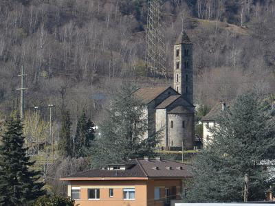 A stone church with a tall, square bell tower and a rounded apse, situated on a hillside behind modern residential buildings and a large electrical pylon.