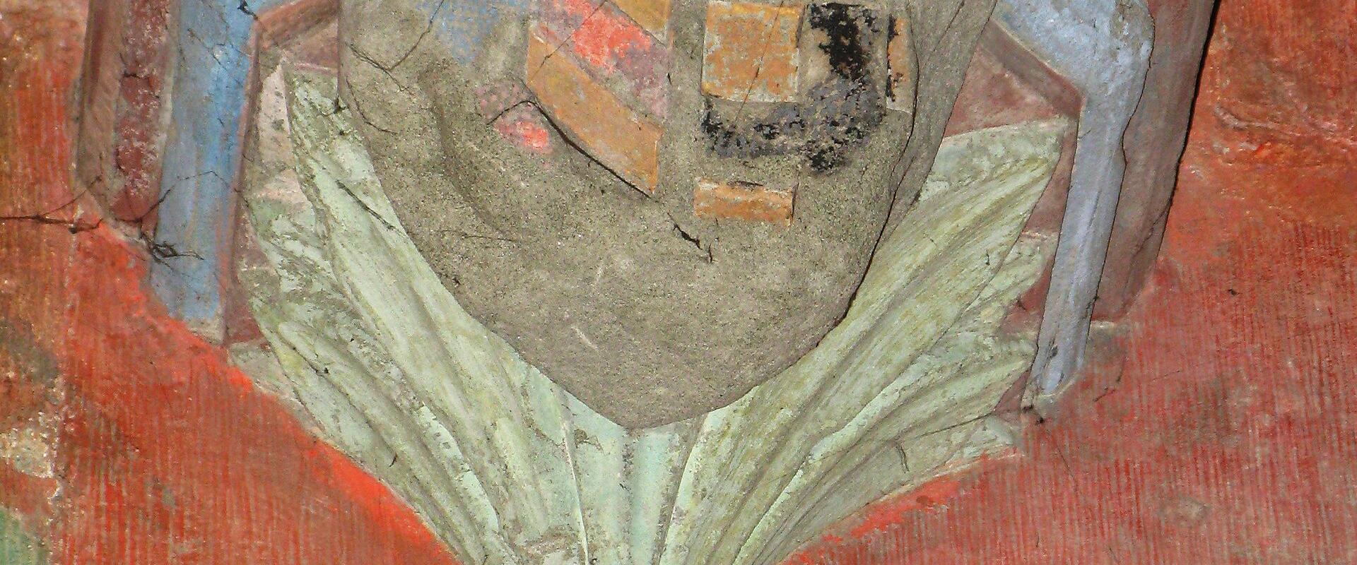 A close-up photograph of a painted stone architectural detail, specifically a column capital featuring a shield with heraldic symbols (a cross and checkered pattern) resting on stylized foliage, set against a red painted wall.