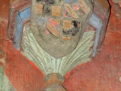 A close-up photograph of a painted stone architectural detail, specifically a column capital featuring a shield with heraldic symbols (a cross and checkered pattern) resting on stylized foliage, set against a red painted wall.