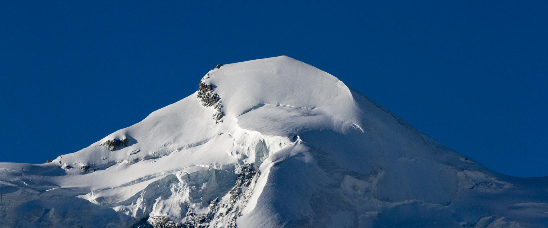 A close-up view of a snow-covered mountain peak with exposed rock faces against a clear blue sky.