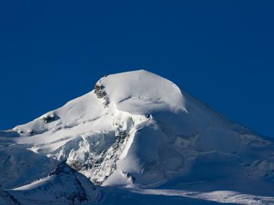 A close-up view of a snow-covered mountain peak with exposed rock faces against a clear blue sky.