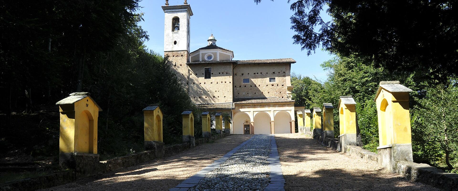 A long, straight processional path lined with yellow chapels leads up to a stone church with a bell tower and a small dome, set against a backdrop of trees and a blue sky.